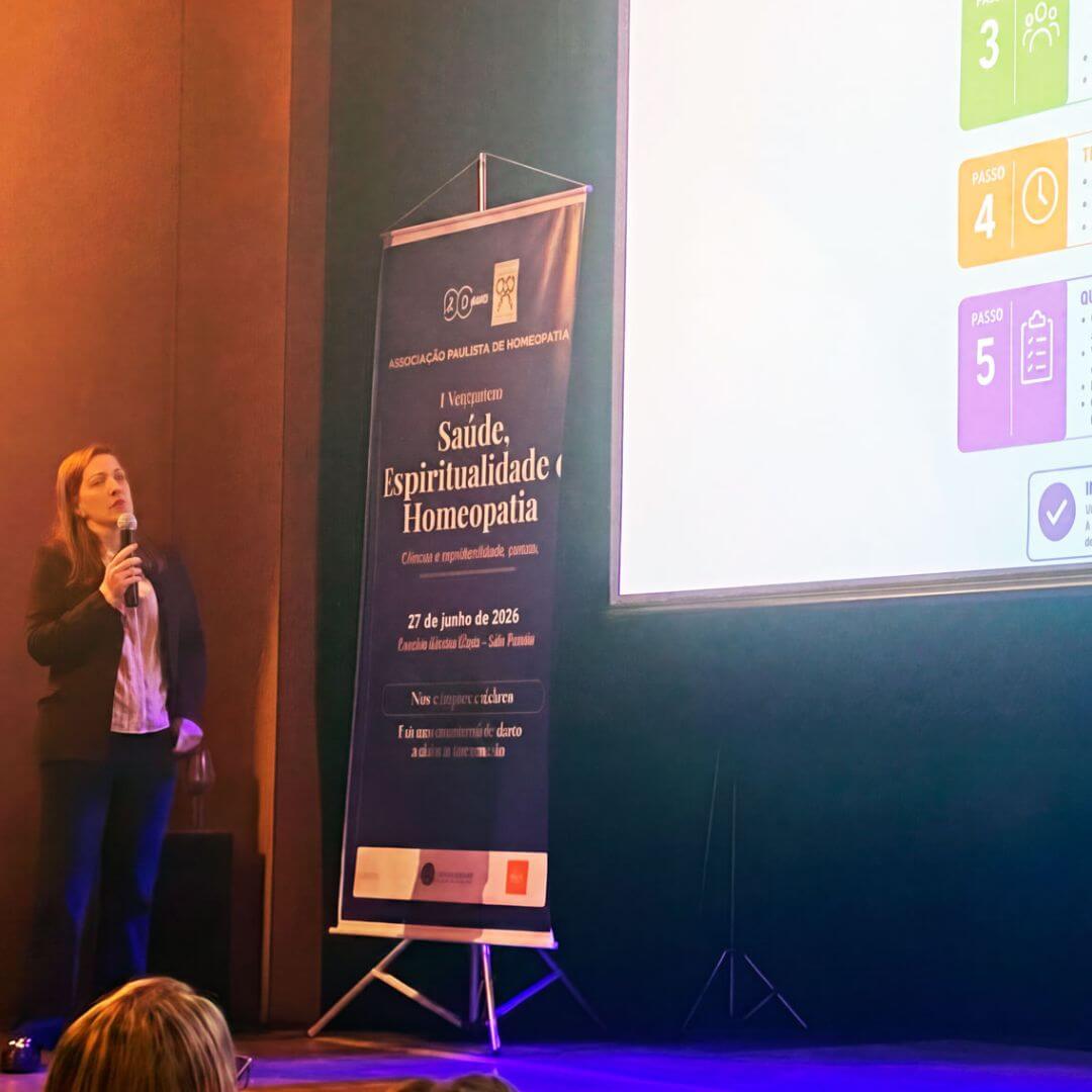 Dra. Camila Casaletti Braghetta durante sua fala no palco do simpósio, ao lado do banner do evento.